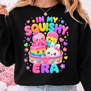 Könnte beinhalten: Ein schwarzes Sweatshirt mit einer bunten Grafik und dem Text IN MY SQUISHY ERA. Das Motiv zeigt einen rosa Oktopus, einen Regenbogenwürfel, ein Eis am Stiel und einen weißen Hasen in einem rosa Korb, umgeben von kleinen Sternen, Herzen und Blumen.