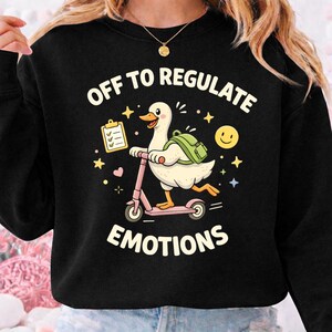 Puede incluir: Sudadera negra con el texto "OFF TO REGULATE EMOTIONS". El gráfico presenta un pato de dibujos animados montado en un patinete rosa, con una mochila y una carpeta. Estrellas, corazones y una carita sonriente rodean al pato.