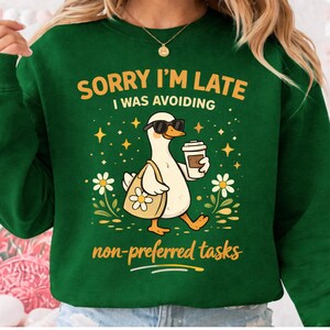 May include: Green sweatshirt with the text "SORRY I'M LATE I WAS AVOIDING non-preferred tasks." The graphic features a white duck wearing sunglasses and carrying a tote bag and coffee. The duck is walking through flowers and stars.