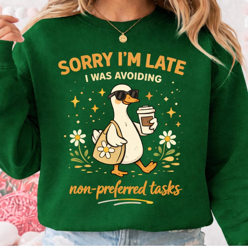 May include: Green sweatshirt with the text "SORRY I'M LATE I WAS AVOIDING non-preferred tasks." The graphic features a white duck wearing sunglasses and carrying a tote bag and coffee. The duck is walking through flowers and stars.