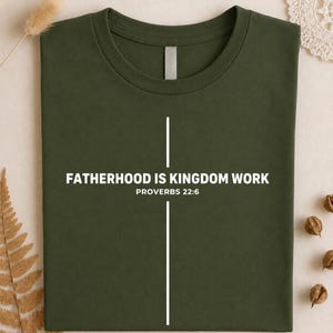 Peut inclure: T-shirt vert olive avec l'inscription blanche &laquo; FATHERHOOD IS KINGDOM WORK &raquo; et &laquo; PROVERBES 22:6 &raquo;. Un motif de croix blanche est &eacute;galement pr&eacute;sent. Le t-shirt est pli&eacute; et pr&eacute;sent&eacute; sur un fond neutre.