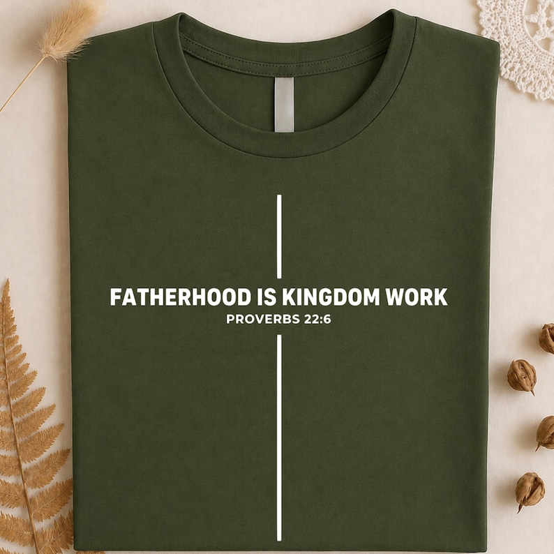 Peut inclure: T-shirt vert olive avec l'inscription blanche &laquo; FATHERHOOD IS KINGDOM WORK &raquo; et &laquo; PROVERBES 22:6 &raquo;. Un motif de croix blanche est &eacute;galement pr&eacute;sent. Le t-shirt est pli&eacute; et pr&eacute;sent&eacute; sur un fond neutre.