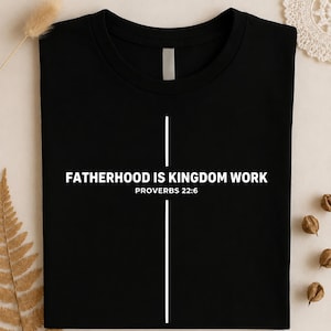 以下が含まれることがあります： 黒いTシャツ。白い文字で「FATHERHOOD IS KINGDOM WORK」と「PROVERBS 22:6」と書かれています。白い縦線がテキストを横切り、上部に短い横線があります。Tシャツはニュートラルな背景に表示されています。