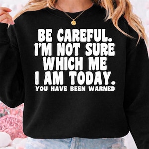 以下が含まれることがあります： 「BE CAREFUL. I'M NOT SURE WHICH ME I AM TODAY. YOU HAVE BEEN WARNED.」という白い文字がプリントされた黒いスウェットシャツ。テキストは太字のサンセリフ体です。スウェットシャツはクルーネックと長袖です。