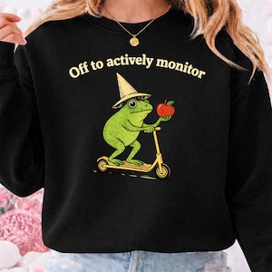 May include: Black sweatshirt with the text "Off to actively monitor." The graphic features a green frog wearing a hat and holding a red apple while riding a yellow scooter. The design is whimsical and playful.