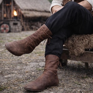 May include: Brown leather boots with a laced upper and a tie closure. The boots have a vintage, worn look, and are designed to reach the mid-calf. The boots are shown with black pants and a rustic background.