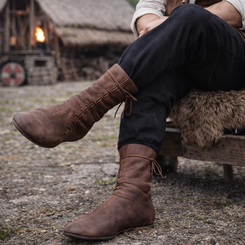 May include: Brown leather boots with a laced upper and a tie closure. The boots have a vintage, worn look, and are designed to reach the mid-calf. The boots are shown with black pants and a rustic background.