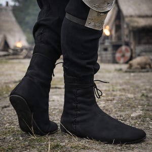 May include: Black suede-like boots with a pointed toe and lace-up detailing. The boots are mid-calf height and appear to be worn in an outdoor setting. The boots are part of a historical or fantasy costume.