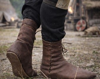 Könnte beinhalten: Braune mittelalterliche Lederstiefel mit Schnürsenkeln. Die Stiefel werden mit einer schwarzen Hose getragen. Die Stiefel haben eine spitze Zehenpartie und sind vorne geschnürt. Die Stiefel sind aus weichem, abgenutztem Leder.