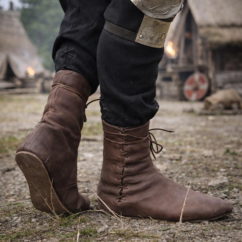 May include: Brown leather medieval boots with laces. The boots are worn with black pants. The boots have a pointed toe and are laced up the front. The boots are made of a soft, worn leather.
