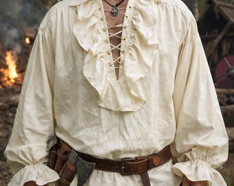May include: Cream-colored pirate shirt with ruffled collar and cuffs. The shirt features a laced neckline and long sleeves. A brown leather belt with a buckle and a small axe are also visible.