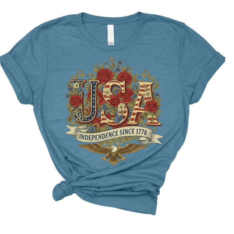 May include: A light blue t-shirt with the word "USA" in a floral design, incorporating red roses and the American flag pattern. The text "INDEPENDENCE SINCE 1776" is on a banner below "USA", with an eagle graphic at the bottom.
