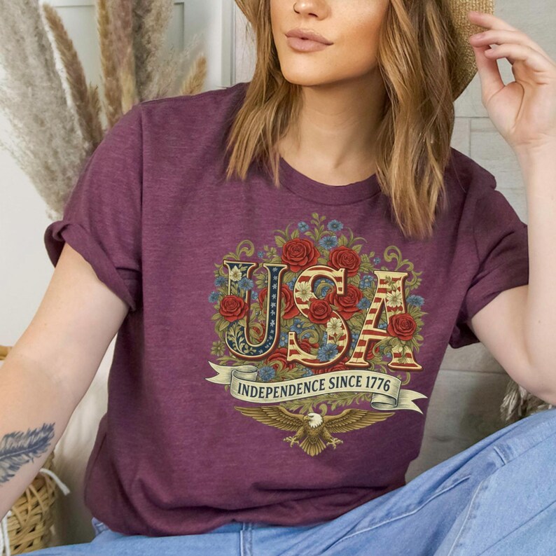 May include: Burgundy t-shirt with the word "USA" in a floral design with red roses and blue flowers. The shirt includes a banner that reads "Independence Since 1776" and an eagle graphic.