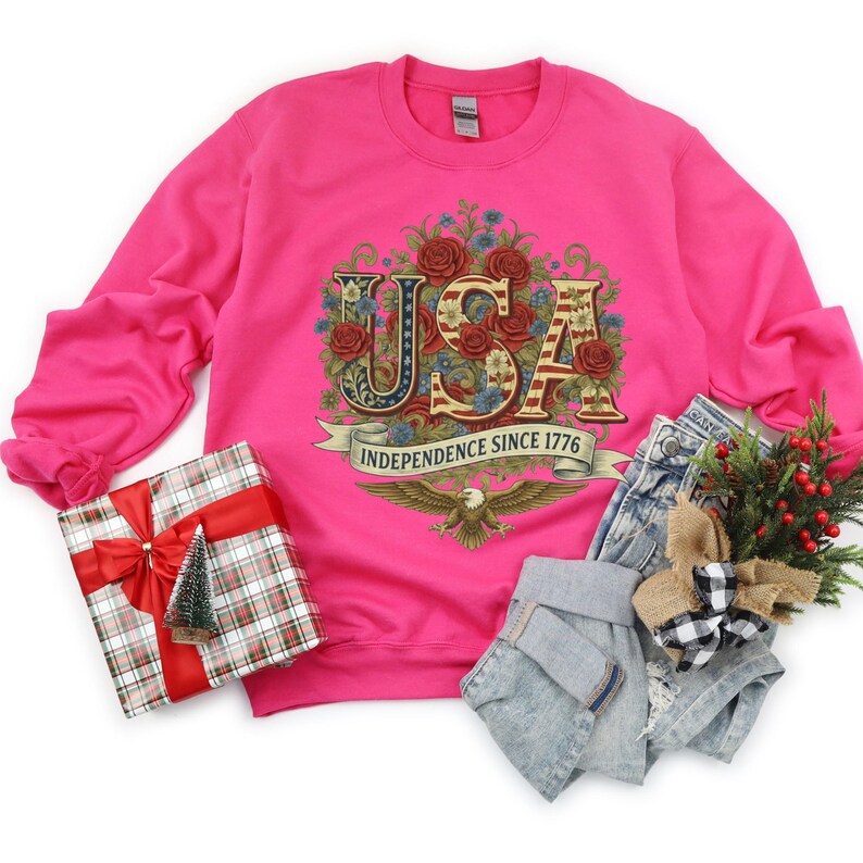 May include: A bright pink sweatshirt featuring the word "USA" in a patriotic design, with floral accents and an eagle. The text "Independence Since 1776" is on a banner. The image also shows a wrapped gift, denim shorts, and festive decorations.