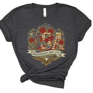 May include: Dark grey t-shirt with a floral "USA" design in red, white, and blue. The text "INDEPENDENCE SINCE 1776" is on a banner, with an eagle below. The design is centrally placed on the shirt.
