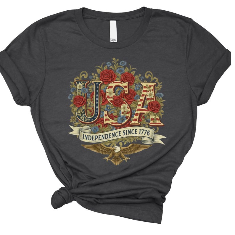 May include: Dark grey t-shirt with a floral "USA" design in red, white, and blue. The text "INDEPENDENCE SINCE 1776" is on a banner, with an eagle below. The design is centrally placed on the shirt.