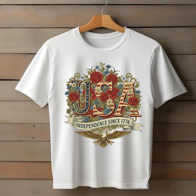 May include: White t-shirt with a patriotic "USA" design, incorporating red, white, and blue colours, roses, and foliage. A banner below reads "Independence Since 1776" with an eagle emblem.