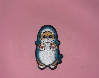 Puede incluir: Parche bordado con un personaje de dibujos animados con un atuendo con capucha azul con vientre blanco y cara naranja. El parche tiene detalles dorados y verde azulado, con pequeños detalles rojos en las manos.