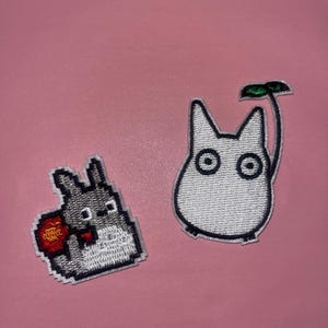 May include: Two embroidered patches featuring characters from the animated film "My Neighbor Totoro." One patch depicts a gray Totoro holding a red object, and the other shows a white character with large eyes and a green leaf.
