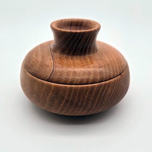 Puede incluir: Un pequeño recipiente de madera redondo con tapa y acabado liso y pulido. El objeto presenta una abertura de cuello acampanado y un patrón de veta de madera visible en tonos marrón medio cálidos, con un ajuste preciso entre la tapa y la base.
