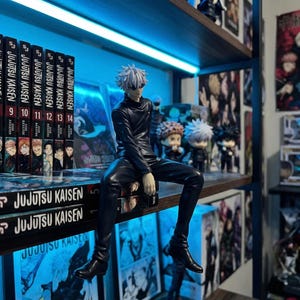 May include: A black figurine of a character from Jujutsu Kaisen, seated on a shelf with manga volumes. The shelf is illuminated with blue light. Other figurines and manga are visible in the background.