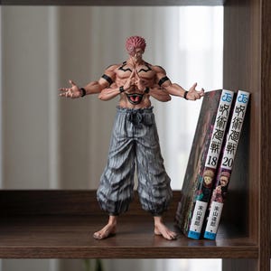 以下が含まれることがあります： 赤い髪と複数の腕を持つキャラクターの精巧なフィギュアで、グレーのズボンを履いて棚に立っています。このフィギュアには複雑なタトゥーがあり、両手を広げています。2冊の日本の漫画本が横に立てかけられています。