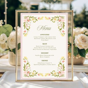May include: A framed menu card with floral accents sits on a wooden table, surrounded by white roses and glassware. The menu features elegant script and a soft pink border, ideal for a wedding or special event. The background includes a blurred outdoor setting.