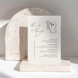 Könnte beinhalten: Eine rechteckige weiße Menükarte mit der Überschrift Boo Bar und einer schwarzen Strichzeichnung eines Geistes, der ein Cocktailglas hält. Der Text listet vier Getränkenamen auf: Ghost-tini, Bloody Mary, Ghostbuster und Ghouls Guts Shot.