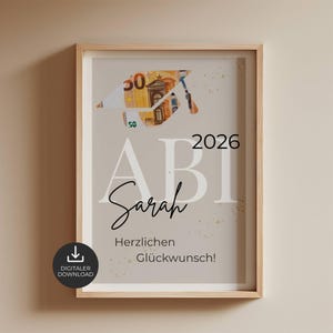 Peut inclure: Une impression encadrée montrant une toque de diplômé pliée à partir d'un billet de 50 euros. Le texte indique ABI 2026, Sarah et Herzlichen Glückwunsch! Le design présente des taches dorées sur un fond beige clair, dans un cadre en bois clair.