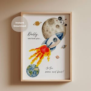 Könnte beinhalten: Ein gerahmter Druck mit einer Aquarellillustration einer Rakete, eines Mondes, Planeten und eines Astronauten. Der Raketenausstoß ist ein Handabdruck. Der Text lautet "Daddy, we love you... to the moon and back!"