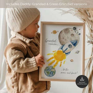 May include: A framed print featuring a watercolor space theme with a rocket, moon, planets, and an astronaut. The design includes a yellow handprint as the rocket's exhaust and the text: Grandad, I love you... to the moon and back!