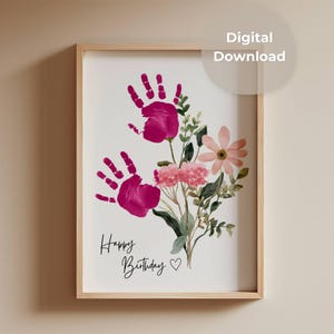 Peut inclure: Une impression numérique encadrée présentant deux empreintes de mains magenta disposées pour ressembler à des fleurs, accompagnées de feuilles vertes et de fleurs roses style aquarelle. Le texte "Happy Birthday" et un cœur sont imprimés en noir.