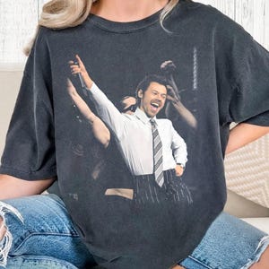 Op de afbeelding: Donkergrijze t-shirt met een concertafbeelding van een man in een wit overhemd, gestreepte stropdas en krijtstreepbroek, die een microfoon vasthoudt en lacht. De afbeelding is gedrukt op een donkergrijze, oversized t-shirt.