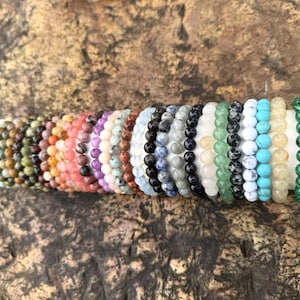 May include: A collection of beaded bracelets in various colors, including brown, orange, pink, purple, blue, black, white, green, and turquoise. The bracelets are displayed on a gold-colored cylindrical stand, showcasing the diverse range of gemstone beads.