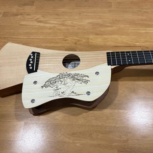 Puede incluir: Una guitarra de viaje de madera clara con un dise&ntilde;o &uacute;nico. La guitarra presenta una boca, cuerdas y una placa decorativa de madera con un dise&ntilde;o grabado de &aacute;rbol y figura. La guitarra descansa sobre una superficie de madera.