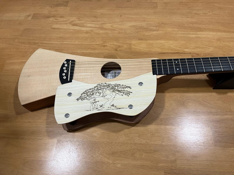 Puede incluir: Una guitarra de viaje de madera clara con un dise&ntilde;o &uacute;nico. La guitarra presenta una boca, cuerdas y una placa decorativa de madera con un dise&ntilde;o grabado de &aacute;rbol y figura. La guitarra descansa sobre una superficie de madera.