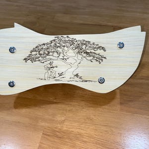 Puede incluir: Una pieza decorativa de madera de color claro con forma curva. Presenta un dise&ntilde;o grabado de un &aacute;rbol con una persona tocando la guitarra debajo. Se ven cuatro sujetadores de color plateado.