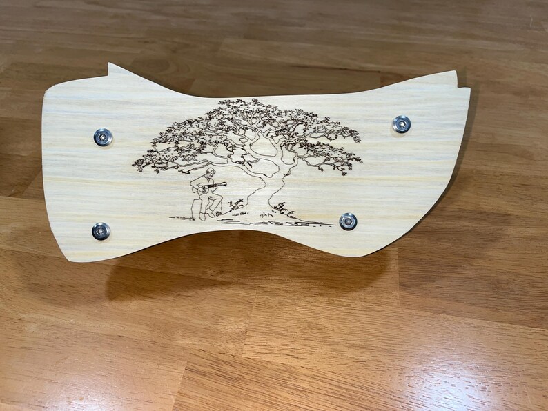 Puede incluir: Una pieza decorativa de madera de color claro con forma curva. Presenta un dise&ntilde;o grabado de un &aacute;rbol con una persona tocando la guitarra debajo. Se ven cuatro sujetadores de color plateado.