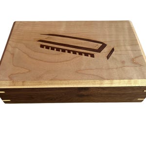 May include: A rectangular wooden box featuring a light-colored wood lid with a dark wood inlay of a harmonica graphic. The sides of the box are constructed from a contrasting dark wood with visible light-colored wood corner joinery details.