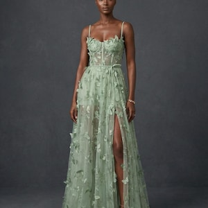 May include: A light green, floor-length gown with butterfly appliqués. The dress features a fitted bodice, spaghetti straps, and a high slit. A thin belt cinches the waist. The model is wearing the dress in front of a dark gray background.