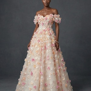 May include: An off-the-shoulder gown with a full skirt, decorated with delicate, three-dimensional floral appliqués in shades of pink, peach, and white. The dress has a fitted bodice and a flowing skirt, ideal for a special event.