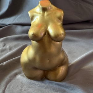 May include: A gold-colored, torso-shaped sculpture. The sculpture is a stylized representation of a human form, with prominent breasts and a rounded lower body. The surface has a smooth, metallic finish.