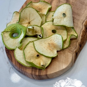 May include: A pile of dried apple slices with green edges and a light brown center, arranged on a wooden cutting board. The board has a natural wood grain pattern. The image also includes a logo that says "Dried Harvest Co."