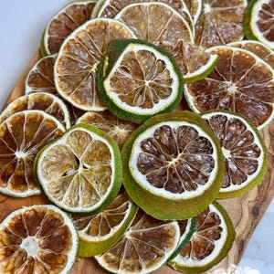 May include: A collection of dried lime slices arranged on a wooden cutting board. The slices have a brown interior with a green rind. The image includes the text "Dried Harvest Co."