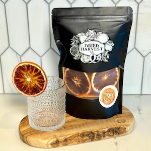 May include: A clear textured glass and a dried blood orange slice rest on a wooden serving board. A black bag with a window displays dried blood orange slices. The bag has a label that reads "Dried Harvest Co." and "dehydrated blood orange."