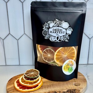 May include: A black bag of dried citrus slices with a clear window, labeled "Dried Harvest Co." and "Variety Pack." The bag sits on a wooden cutting board with a stack of dried citrus slices.