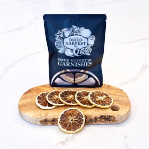 Peut inclure: Un sachet noir de "Dried Harvest Co. Premium Cocktail Garnishes" avec des tranches d'orange séchées sur une planche en bois. Le sachet présente un logo avec des citrons et des herbes. Les tranches d'orange sont disposées devant le sachet.