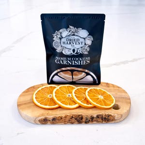 Könnte beinhalten: Ein schwarzer Beutel mit "Dried Harvest Co." Premium-Cocktail-Garnituren steht hinter vier getrockneten Orangenscheiben auf einem Holzbrett. Der Beutel hat eine weiße Illustration von Zitrusfrüchten und Kräutern.