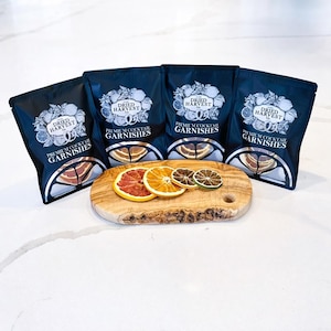 May include: Four black bags of dried cocktail garnishes with the text "Dried Harvest Co. Premium Cocktail Garnishes". A wooden cutting board holds slices of dried grapefruit, orange, and lime. The garnishes are arranged on a white surface.
