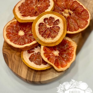 May include: Dried grapefruit slices arranged on a wooden cutting board. The slices are a vibrant red-orange color with a yellow rind. The image includes a logo with the text "Dried Harvest Co."